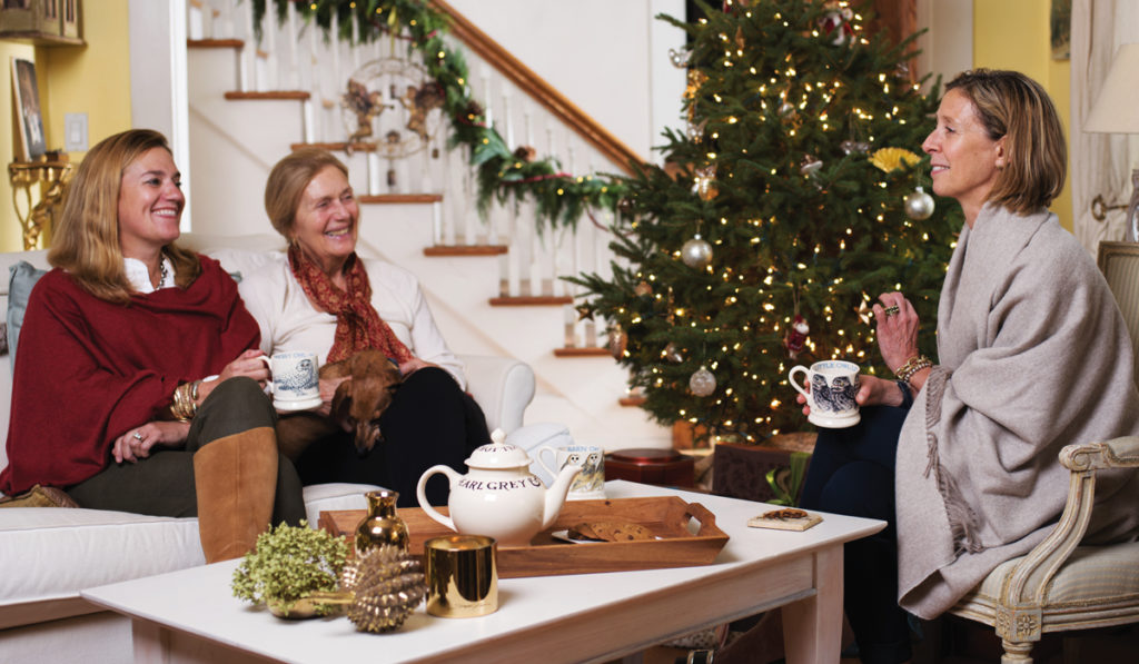 3 women sitting in living room next to Christmas treen drinking tea.